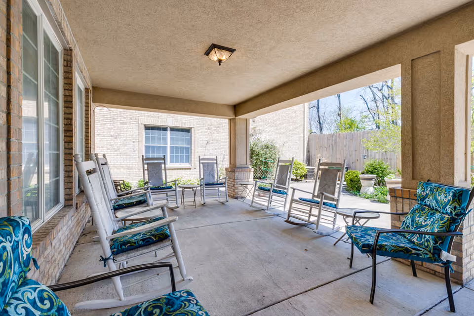 Covered outdoor patio area with several white rocking chairs and cushioned chairs arranged around small tables. The patio is adjacent to a brick building with windows, and there are plants and a wooden fence visible in the background.