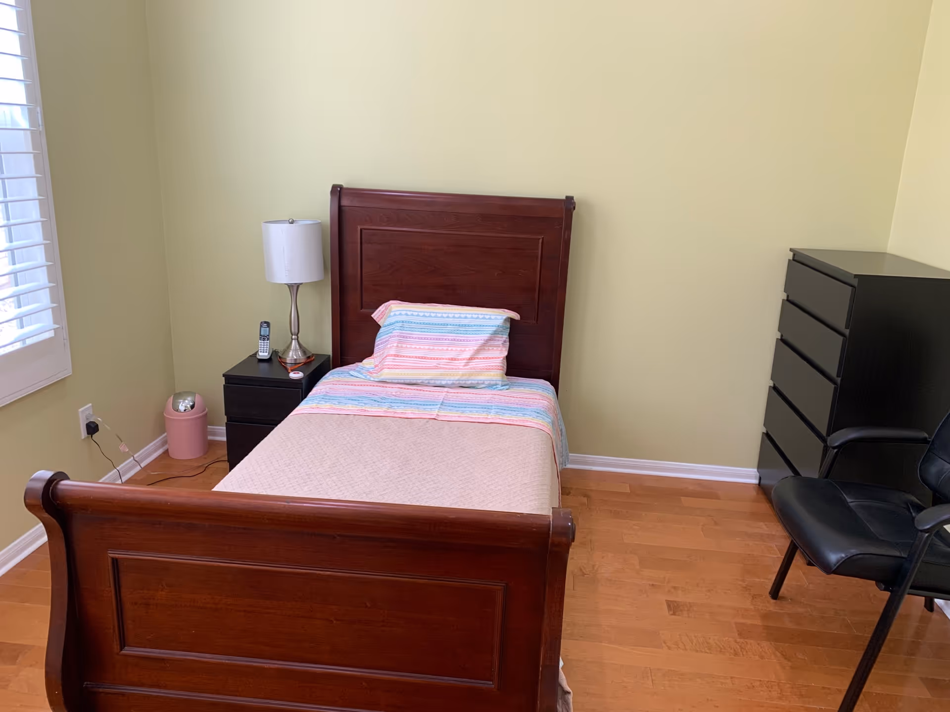 A small bedroom with a single wooden bed that has a colorful striped pillow and beige blanket. Next to the bed is a black nightstand with a silver lamp and a cordless phone. There is a black chest of drawers and a black chair on the right side of the room. The walls are light yellow and the floor is wooden. A window with white blinds is partially visible on the left.