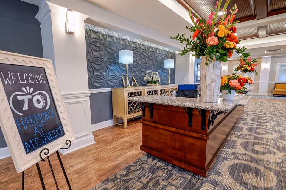 A welcoming lobby at Harmony at Mt. Juliet featuring a reception desk with a granite countertop, decorative floral arrangements, and a chalkboard sign that reads 'Welcome to Harmony at Mt. Juliet'.
