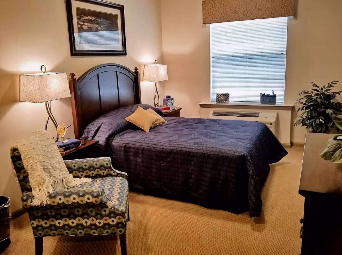 Well-lit furnished bedroom with a dark wood headboard, navy bedspread, patterned armchair, bedside lamps, and a window with blinds.