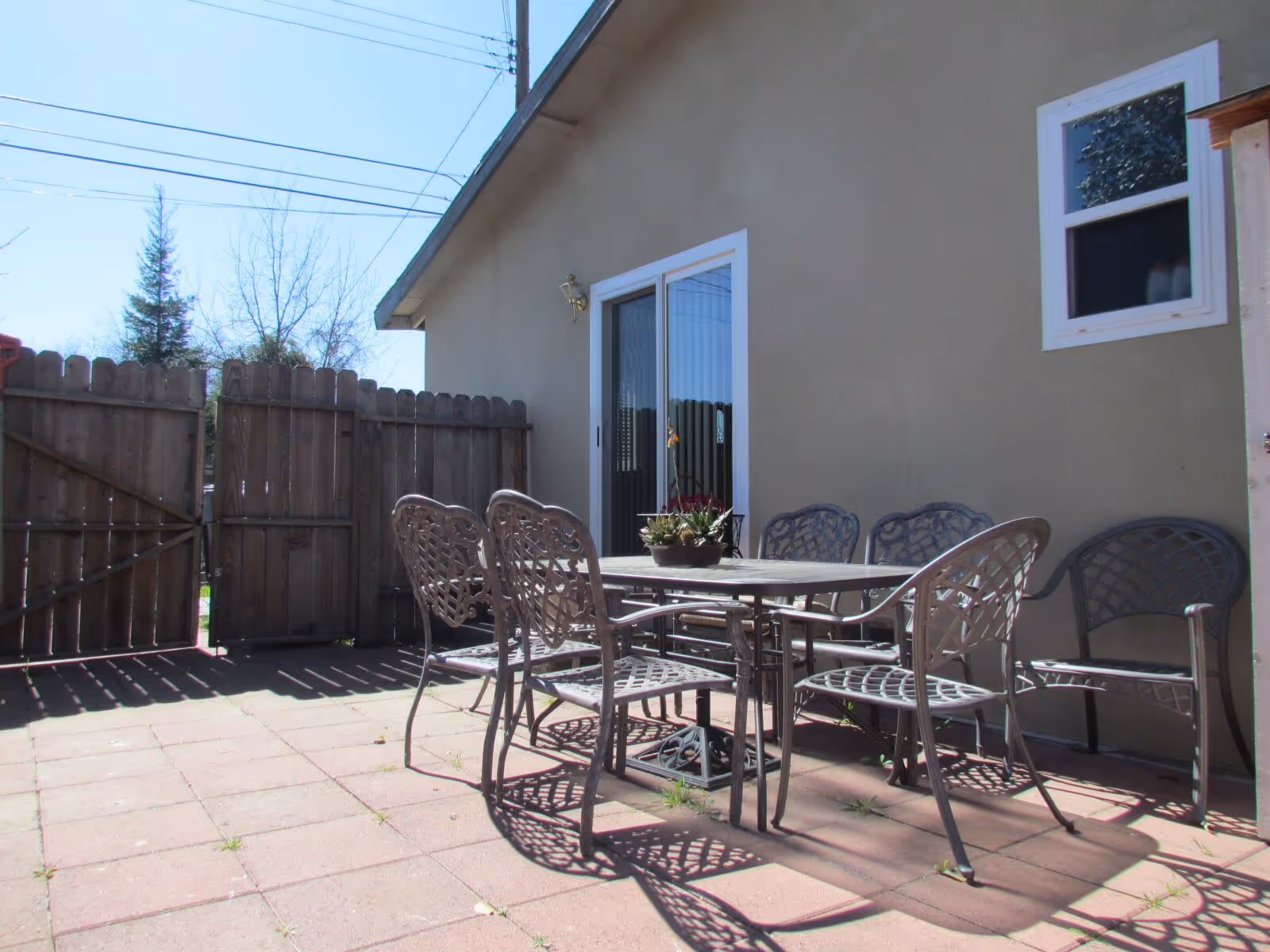 Outdoor patio area with a metal table and six matching chairs on a tiled floor, next to a beige building with a sliding glass door and a small window. A wooden fence encloses the patio, and trees and power lines are visible in the background under a clear blue sky.
