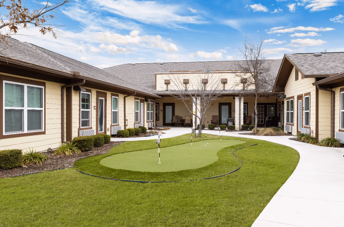 Outdoor courtyard area of a senior living facility with a small putting green surrounded by a curved concrete walkway. The building has beige siding with brown trim and multiple windows and doors facing the courtyard. There are some leafless trees and shrubs around the area under a partly cloudy sky.