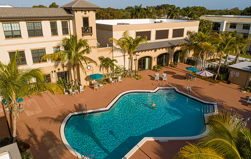 Outdoor swimming pool area at Abbey Delray Health Care Center with lounge chairs, umbrellas, palm trees, and a person swimming in the pool. The building surrounds the pool with multiple windows and doors opening to the pool area.