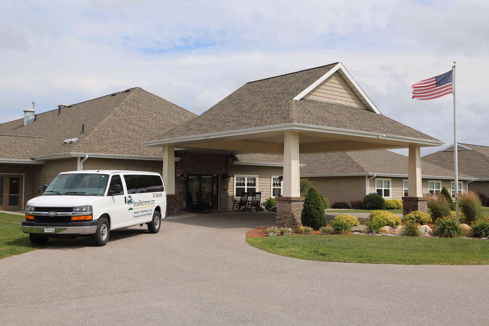 Exterior view of The Brook of Big Rapids senior living facility showing the main entrance with a covered drop-off area, a white van parked nearby with the facility's website and phone number on its side, an American flag on a flagpole, and landscaped greenery around the driveway.