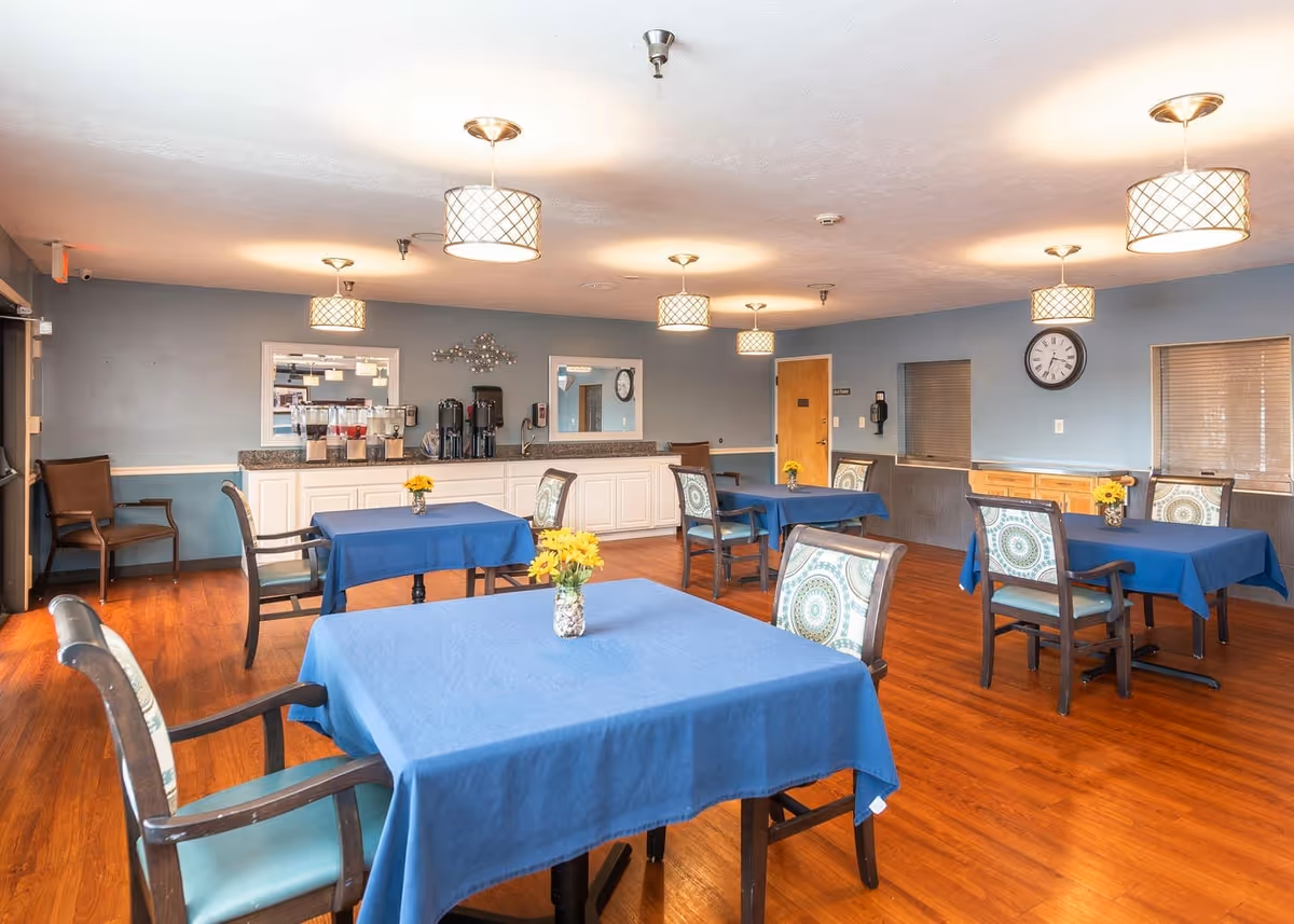 Dining room with several tables covered with blue tablecloths, each table has a small vase with yellow flowers. The room has wooden floors, blue walls, and multiple hanging light fixtures. There is a counter with beverage dispensers and coffee machines against one wall, and a clock on another wall.