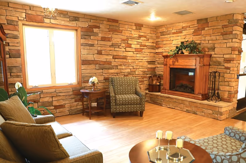A cozy living room with stone walls, a wooden floor, and a wooden fireplace with a plant on top. The room has a window letting in natural light, a patterned armchair, a sofa with cushions, a round wooden coffee table with candles, and a small side table with a flower vase.