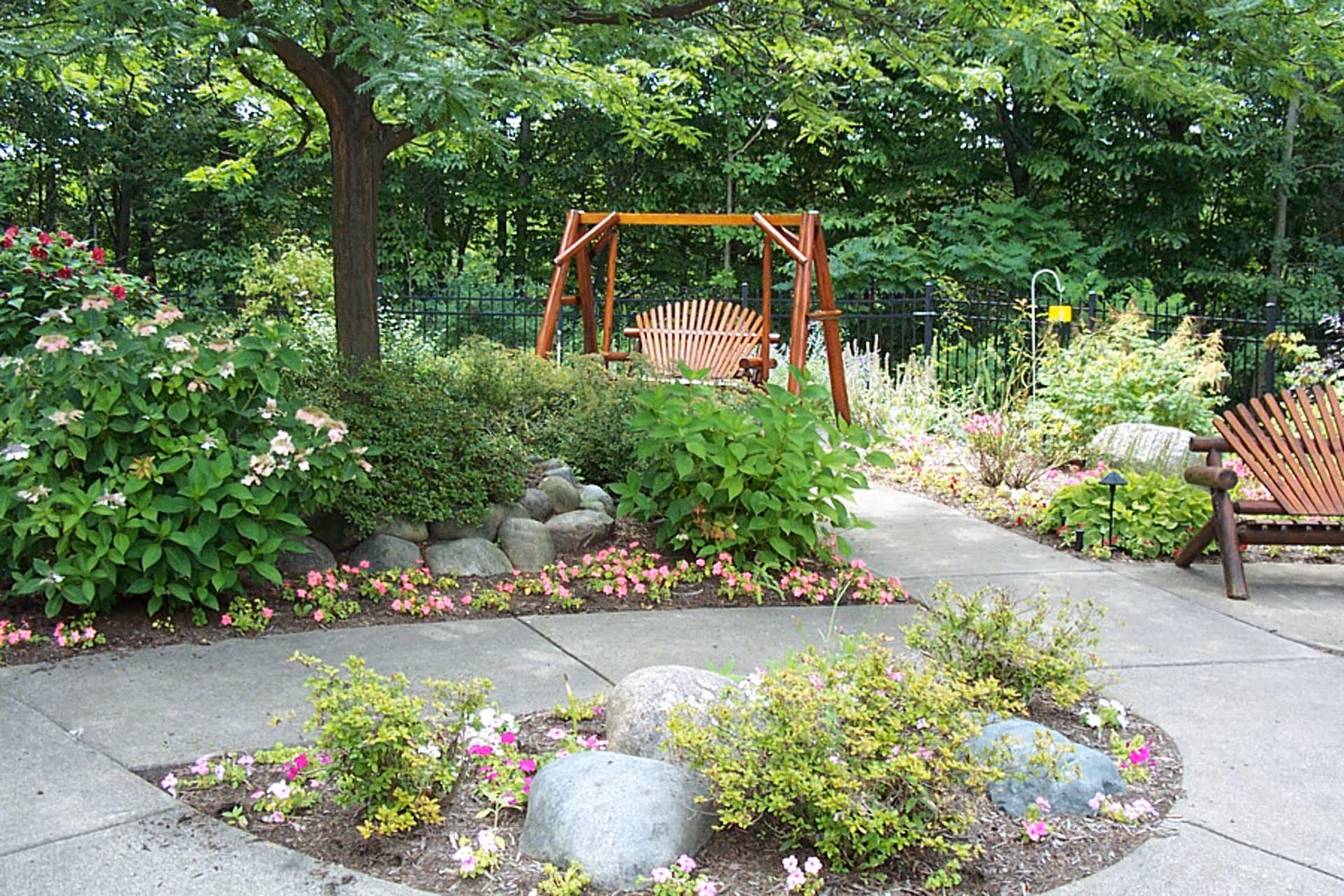 A peaceful outdoor garden area with a concrete pathway surrounded by various green plants, bushes, and colorful flowers. There is a wooden swing bench and a wooden chair placed on the right side, with a backdrop of tall trees and a black metal fence.