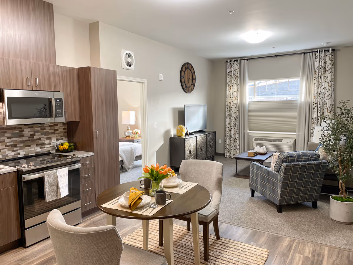 A bright and modern living space in a retirement community featuring a small round dining table set for two with plates, mugs, and a vase of orange tulips. To the left is a kitchen area with wood cabinetry, a microwave, stove, and a tiled backsplash. In the background, there is a living room area with a plaid armchair, a coffee table, a TV on a decorative cabinet, and a window with floral curtains. A doorway leads to a bedroom with a bedside lamp and flowers visible.