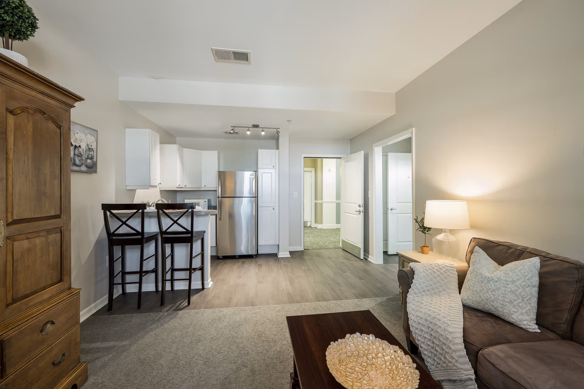 Open-concept living room and kitchenette with a sofa, coffee table, bar stools at a breakfast bar, and a stainless steel refrigerator.