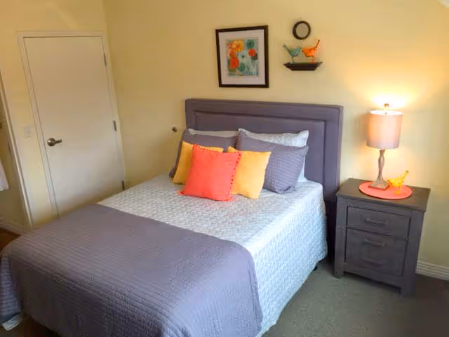 A neatly made bed with a gray quilted bedspread and colorful pillows in shades of orange and yellow. Above the bed is a framed floral painting and two decorative bird figurines on a small wall shelf. To the right of the bed is a dark wooden nightstand with a lamp and a small yellow bird figurine on a red placemat. A closed white door is visible on the left side of the room.