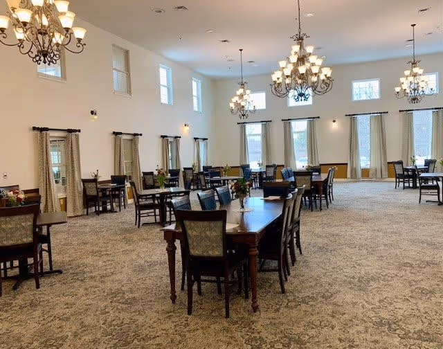 A spacious dining room with multiple wooden tables and chairs arranged neatly. The room has large windows with curtains, high ceilings, and elegant chandeliers providing warm lighting. The carpet has a patterned design, and small flower arrangements are placed on the tables.