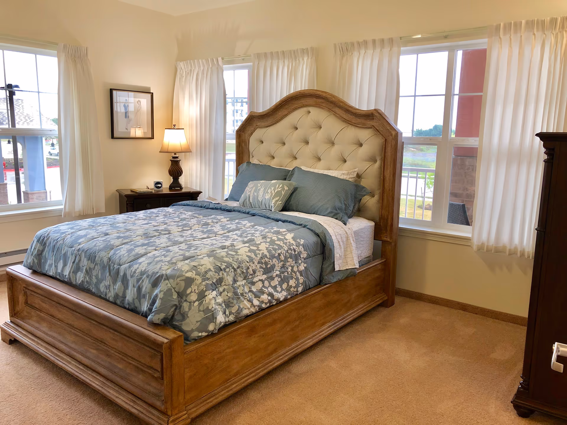Sunlit bedroom with a wooden bed featuring an upholstered headboard, blue floral bedding, a nightstand with a lamp, and windows with white curtains.