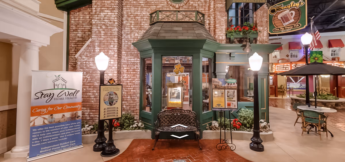 Indoor scene resembling a small town street with a brick wall, green-trimmed windows, and a bench in front. There are two black street lamps, a table with chairs and an umbrella, and various signs including one for 'Stay Well Home Health' and another for 'O'Smyley's One Big Happy Pub'. The setting appears to be a common area designed to look like an outdoor street within a facility.
