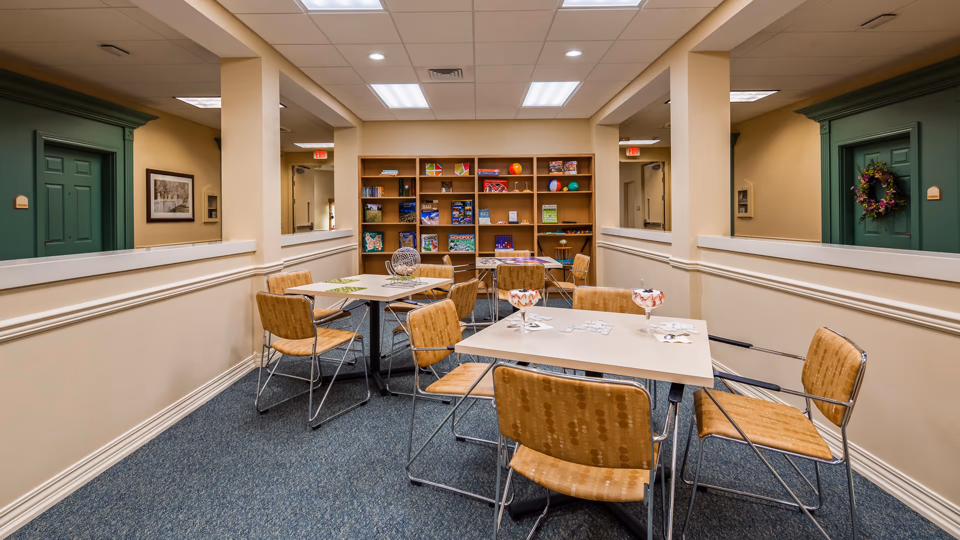 A well-lit common area with tables and chairs arranged for games or activities. The room has beige walls, blue carpet, and a bookshelf filled with board games and puzzles at the far end. There are two green doors on either side of the room, one decorated with a floral wreath. The ceiling has recessed lighting and a drop ceiling design.