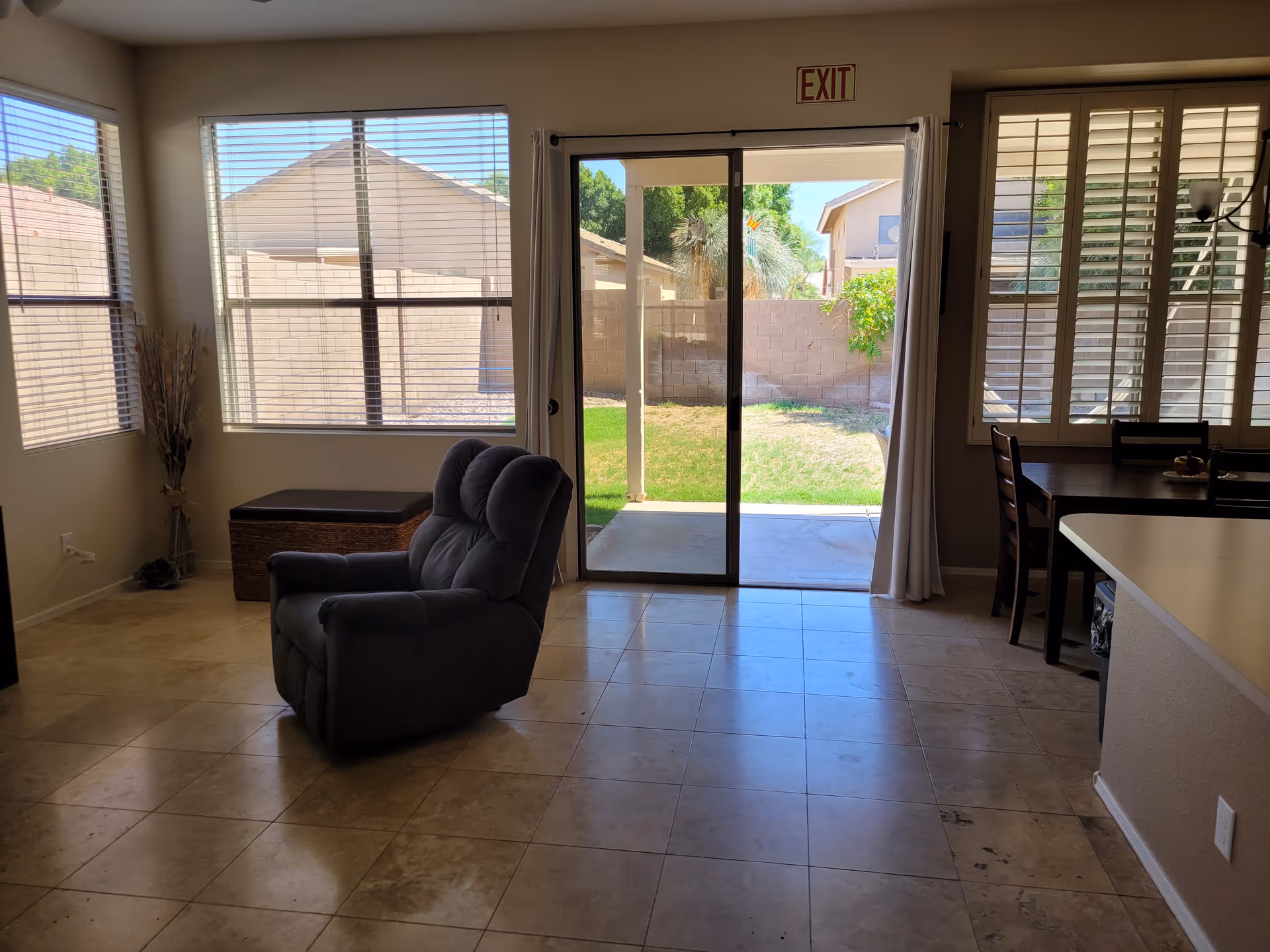 Interior view of a living room with a single gray recliner chair in the center, large windows with blinds on the left and right walls, a sliding glass door leading to a backyard with grass and a concrete patio, and a dining table with chairs on the right side.