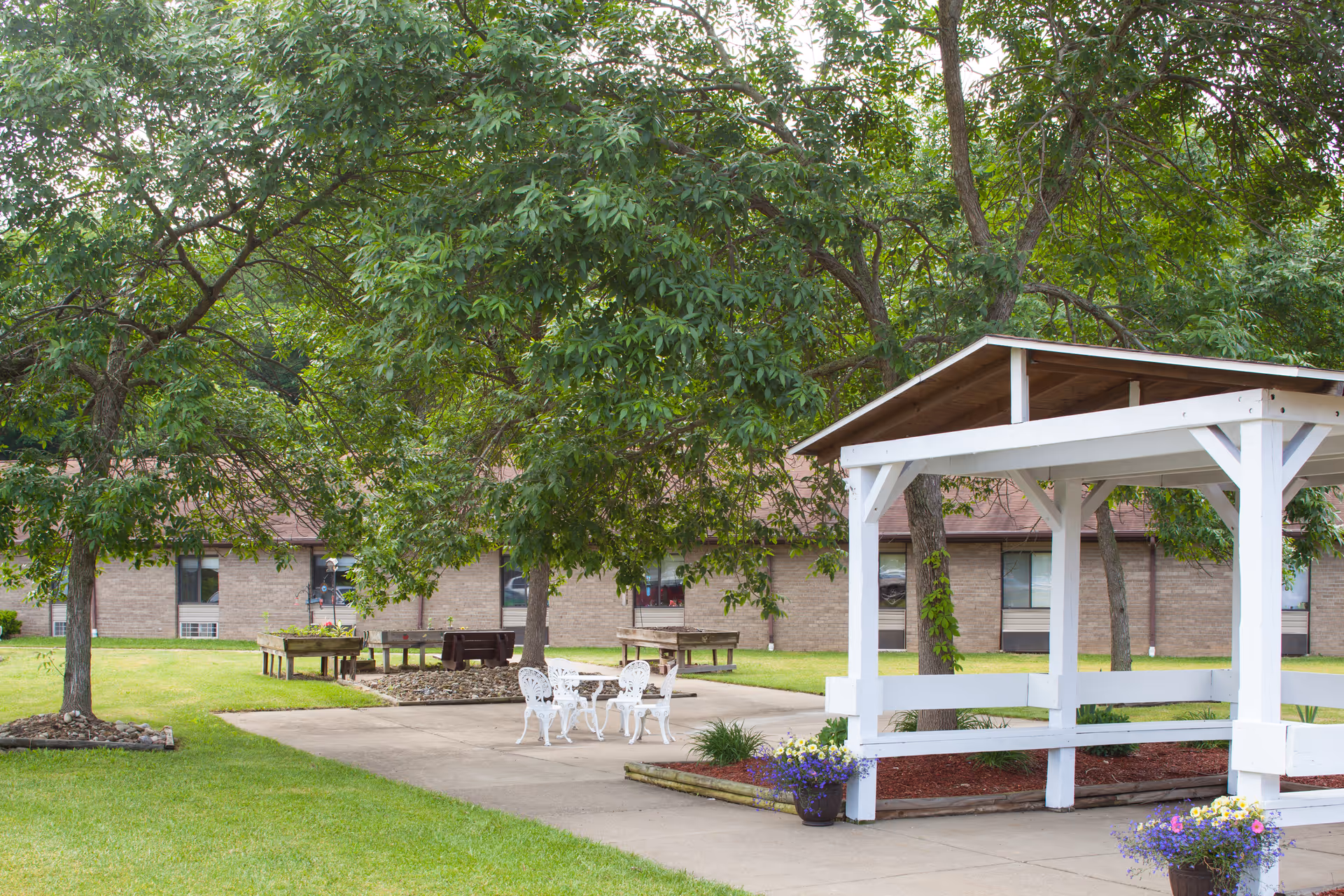 Outdoor garden area at Sistersville Center featuring a white wooden gazebo, white metal table and chairs, several trees, flower pots, and a brick building in the background.
