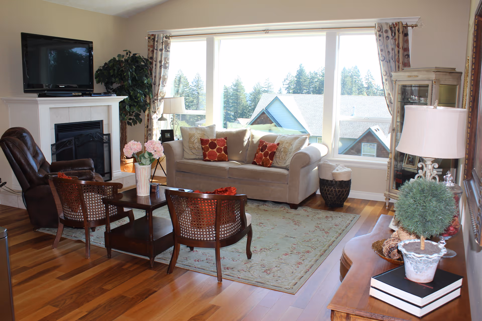 A cozy living room with a beige sofa adorned with decorative pillows, two wooden chairs with woven backs, a dark leather recliner, and a wooden coffee table with a vase of pink flowers. The room features a fireplace with a TV mounted above it, a large window with a view of houses and trees outside, a floor lamp, a glass display cabinet, and a wooden side table with a small potted plant and books.
