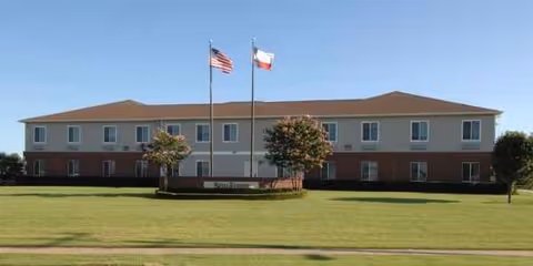 Two flagpoles with American and Texas flags in front of a two-story senior living building.