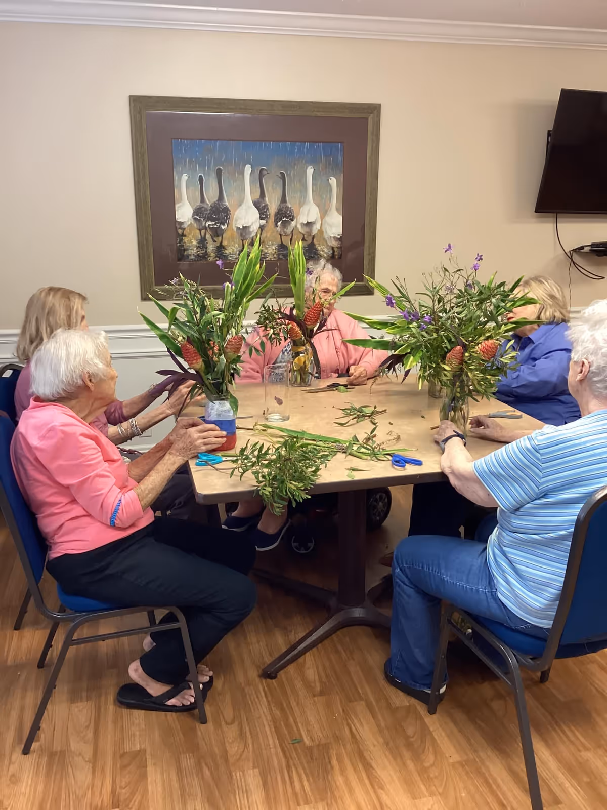 A group of elderly women sitting around a table in a room, arranging flowers in glass jars. The table has scissors and plant clippings scattered on it. A painting of geese is hanging on the wall behind them, and a TV is mounted on the wall to the right.