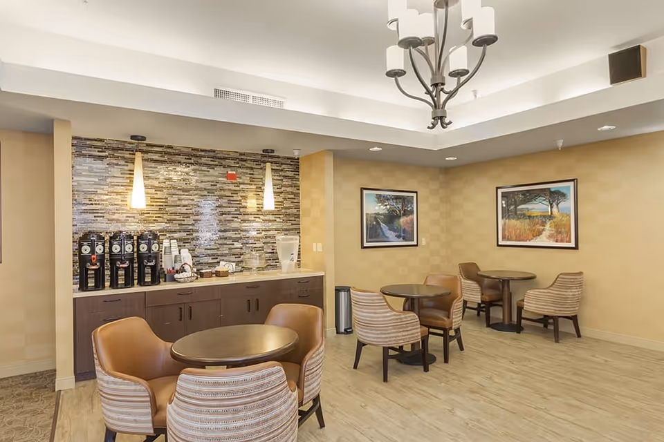 Communal dining and coffee area with round tables, upholstered chairs, and a coffee station against a tiled backsplash.