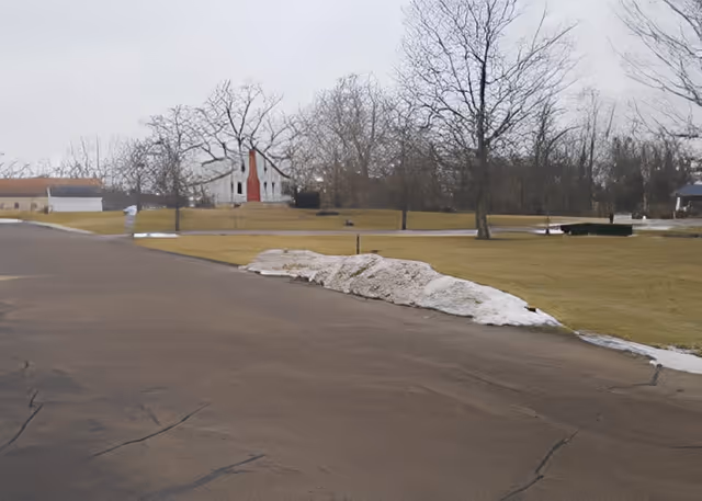 A paved road with patches of melting snow on the side, leading towards a white building with a red door in the background. Leafless trees and a grassy area surround the road under an overcast sky.
