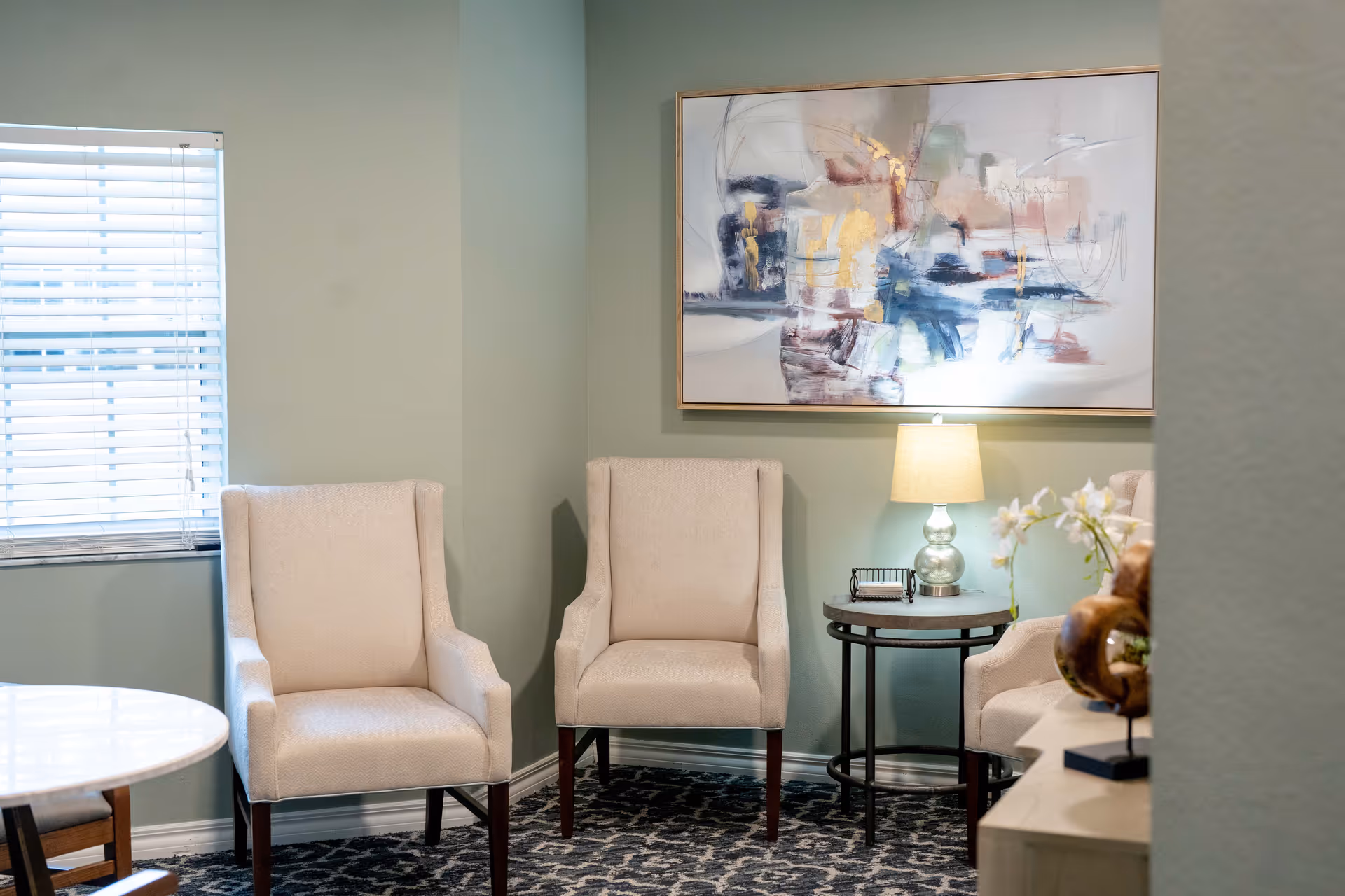 A cozy sitting area with two beige upholstered armchairs placed against a light green wall. Between the chairs is a round side table with a glass lamp and a small decorative item. Above the table hangs an abstract painting with soft colors. A window with white blinds is on the left side, and part of a round white table and another chair are visible in the foreground.