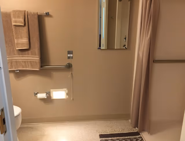A bathroom with beige walls and floor, featuring a toilet partially visible on the left, a towel rack with two brown towels, a grab bar, a toilet paper holder with toilet paper, a small wall-mounted light, a rectangular mirror, and a beige shower curtain on the right side. A small rug is placed on the floor near the shower area.