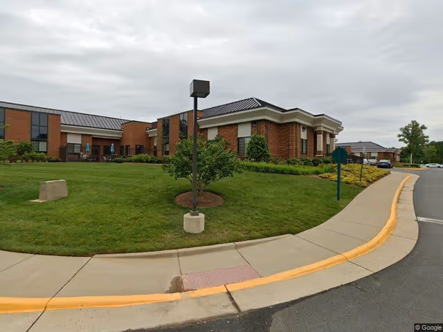 Exterior view of a brick building with a manicured lawn, small trees, and shrubs. The building has a dark roof and multiple windows. A sidewalk curves around the lawn with a streetlamp and a street sign visible. The sky is overcast.