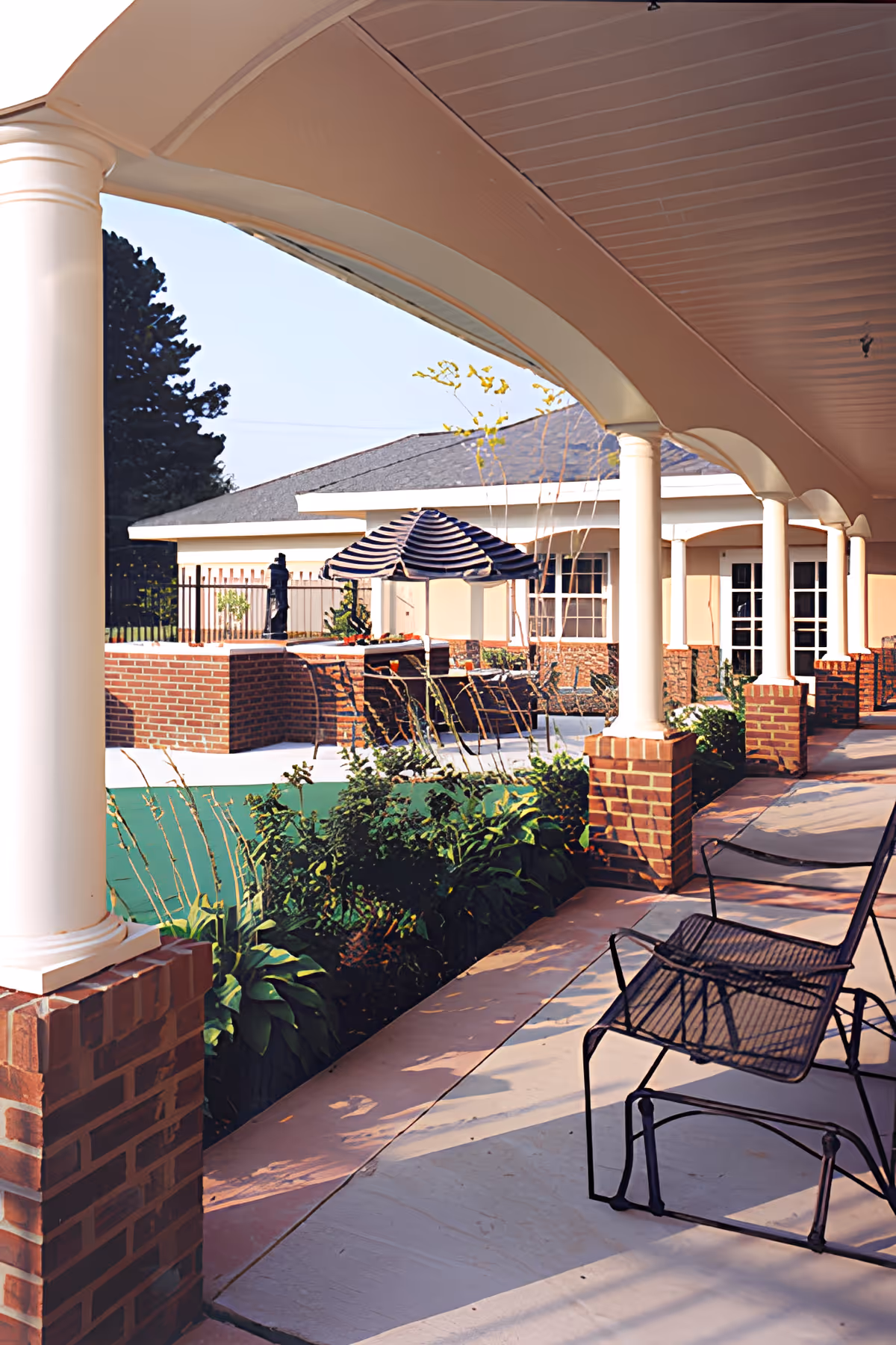 Covered outdoor patio area with white columns and brick bases, metal chairs, and a view of a garden with plants and a striped umbrella over a seating area in the background.