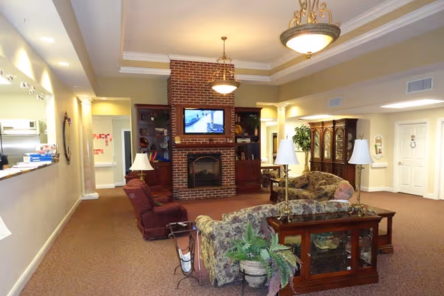 A cozy living room area in a senior living facility featuring a brick fireplace with a mounted TV above it, surrounded by wooden shelves. The room has floral patterned sofas, a burgundy armchair, wooden side tables with lamps, and a potted plant. The walls are painted beige with white trim, and there are two ceiling light fixtures. A man is sitting on one of the sofas, and there is a hallway and a cabinet visible in the background.