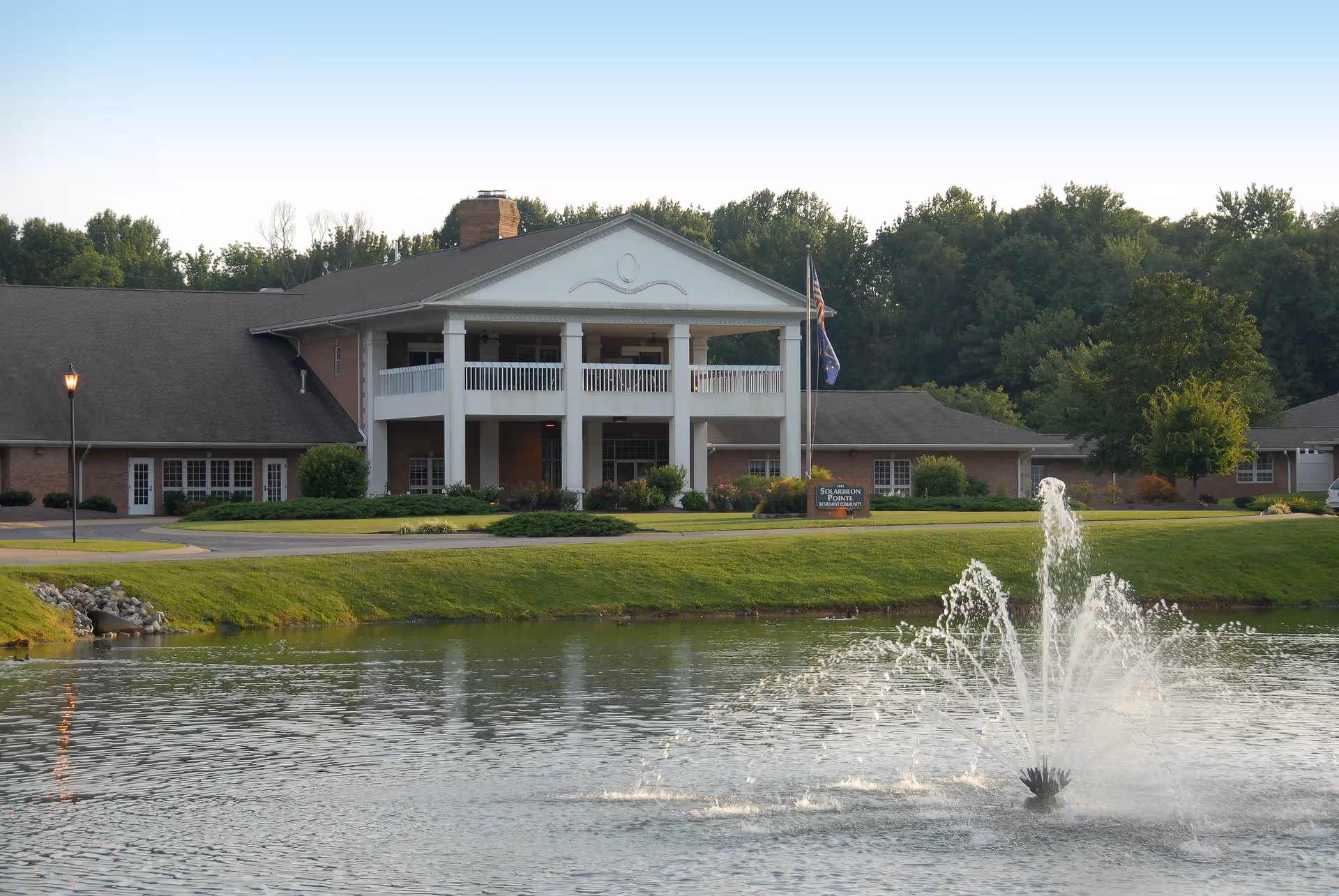 A large senior living facility building with a white columned entrance and balcony, surrounded by greenery and trees. In the foreground, there is a pond with a water fountain spraying water upwards. An American flag and another flag are visible near the entrance.
