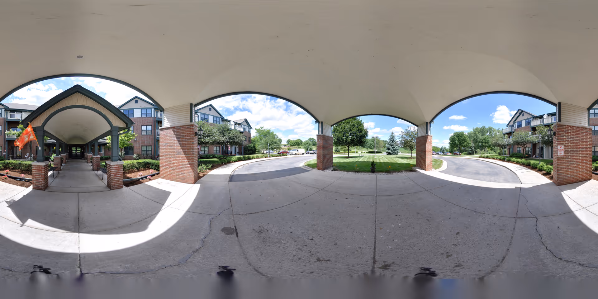 Covered driveway entrance of a senior living facility with brick pillars and arches, leading to a landscaped area with trees and shrubs under a blue sky with scattered clouds.