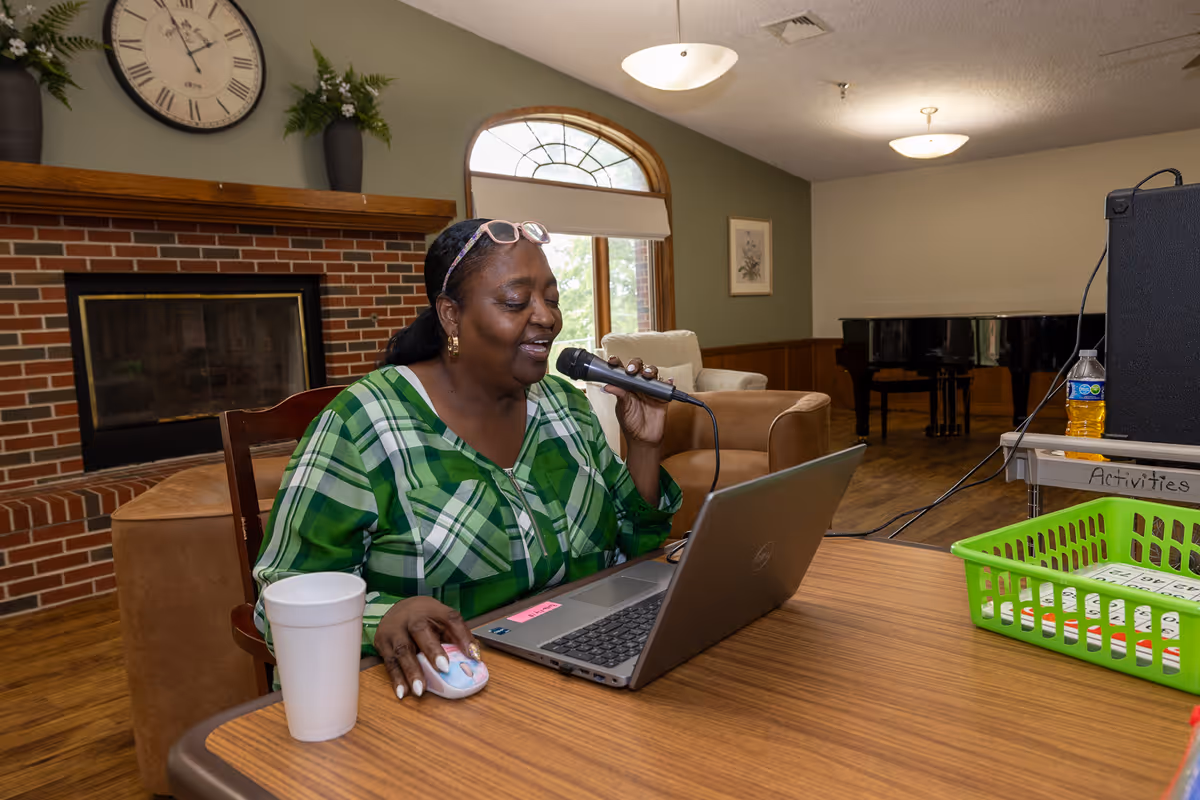 A woman in a green plaid shirt sits at a wooden table in a cozy room with a brick fireplace and a large clock on the wall. She is speaking into a microphone while using a laptop and a computer mouse. The room has a large arched window, comfortable chairs, a piano in the background, and a green basket labeled 'Activities' on the table.