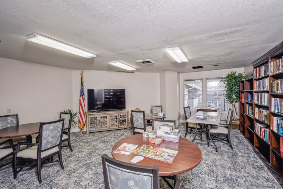 A well-lit common room with multiple round tables and chairs arranged for group activities. There are board games and puzzles on the tables. A large flat-screen TV is mounted on a cabinet against the wall, with an American flag standing next to it. A tall bookshelf filled with books lines one side of the room, and a large window with a potted plant nearby lets in natural light.