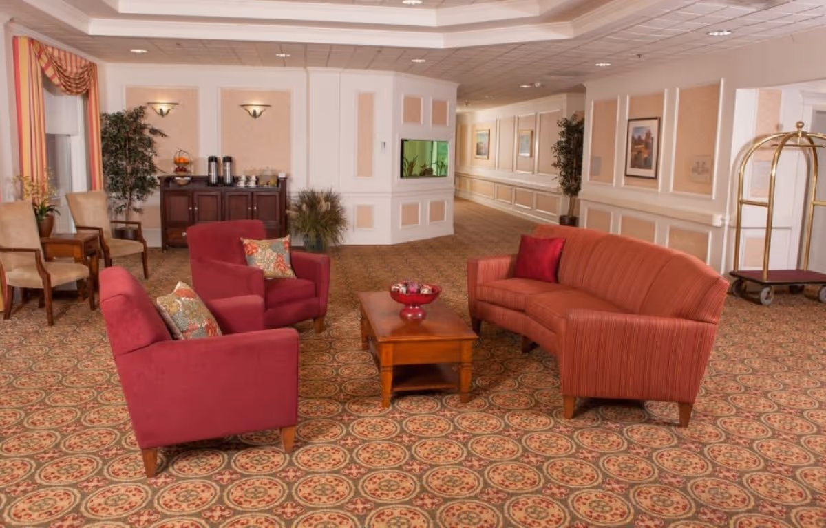 A cozy seating area in a senior living facility with two red armchairs and a red striped sofa arranged around a wooden coffee table. The room features patterned carpet, beige walls with white trim, framed artwork, potted plants, a sideboard with coffee dispensers, and a luggage cart near the hallway.