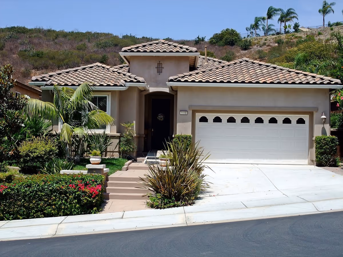 Front exterior view of a single-story house with a tiled roof, a two-car garage with white doors, a small front porch, and landscaped greenery including bushes, palm trees, and flowering plants. The house is situated on a slightly sloped street with a hill and more vegetation in the background under a clear blue sky.