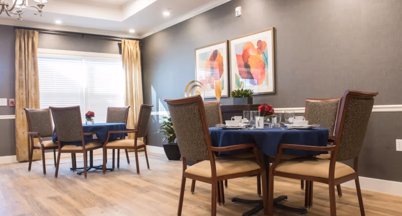 A dining room with two round tables covered with blue tablecloths, each surrounded by four wooden chairs with patterned backs and beige cushions. The tables are set with white dishes, glasses, and silverware. The room has light wood flooring, gray walls with white trim, two colorful abstract paintings, and large windows with beige curtains letting in natural light.