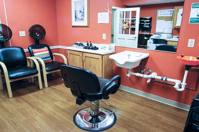 Interior of a small salon or barber area with a black salon chair in the center, two black waiting chairs with wooden armrests and a striped pillow against a red wall, a small white sink mounted on the wall with visible plumbing, a wooden cabinet with items on top, and a mirror reflecting part of the room.