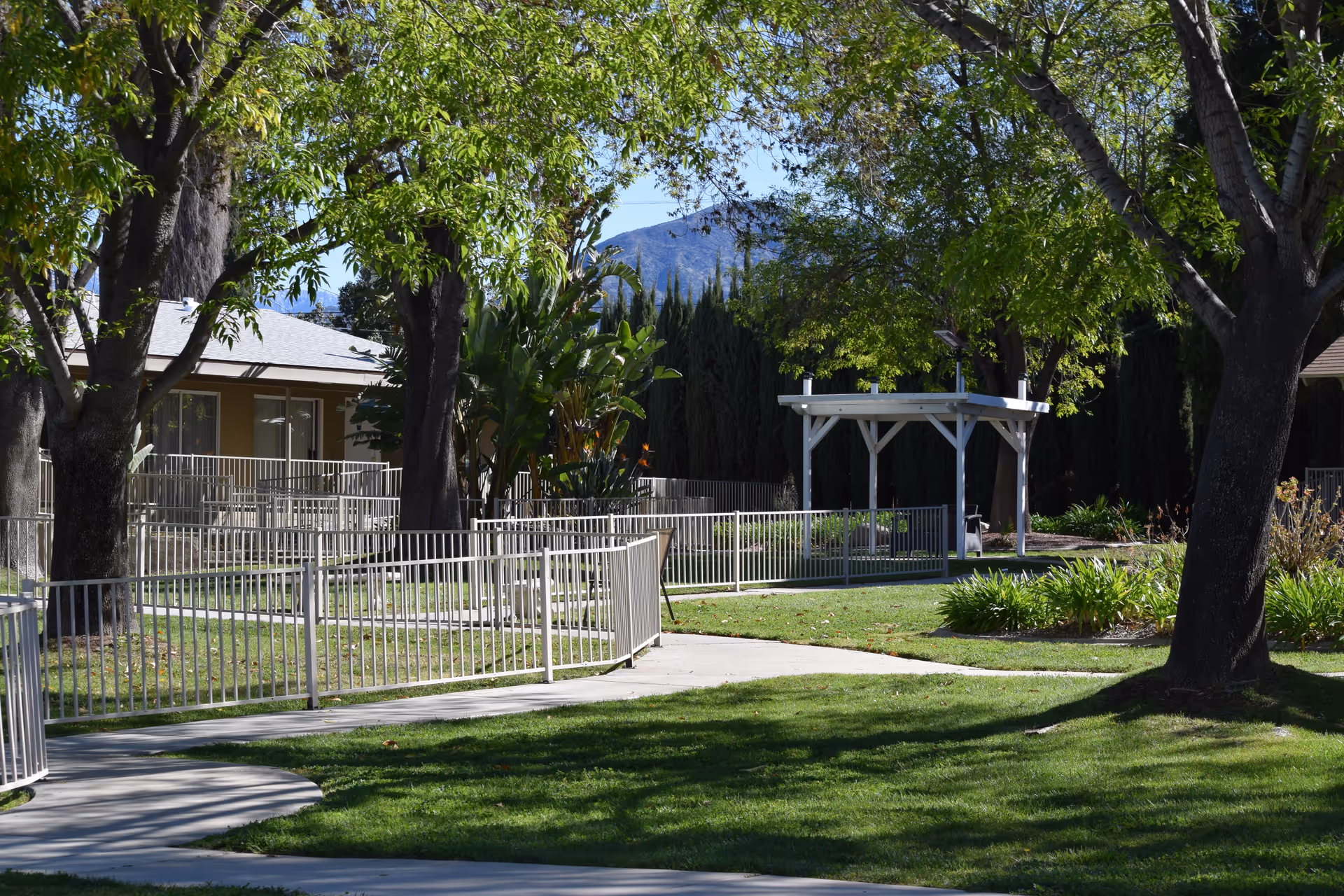 A sunny outdoor garden area with green grass, trees, and a paved walkway with metal railings. There is a small white gazebo structure in the background and a building partially visible on the left side. Mountains can be seen in the distance under a clear blue sky.