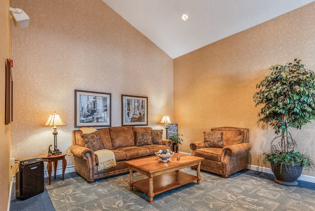A cozy living room with a brown leather sofa and matching armchair, both adorned with decorative pillows. A wooden coffee table sits in front of the sofa with a decorative bowl on top. Two framed black and white pictures hang on the beige patterned wall behind the sofa. Two table lamps with beige shades provide warm lighting. A large potted plant is placed in the corner of the room, and the floor is covered with a patterned carpet.