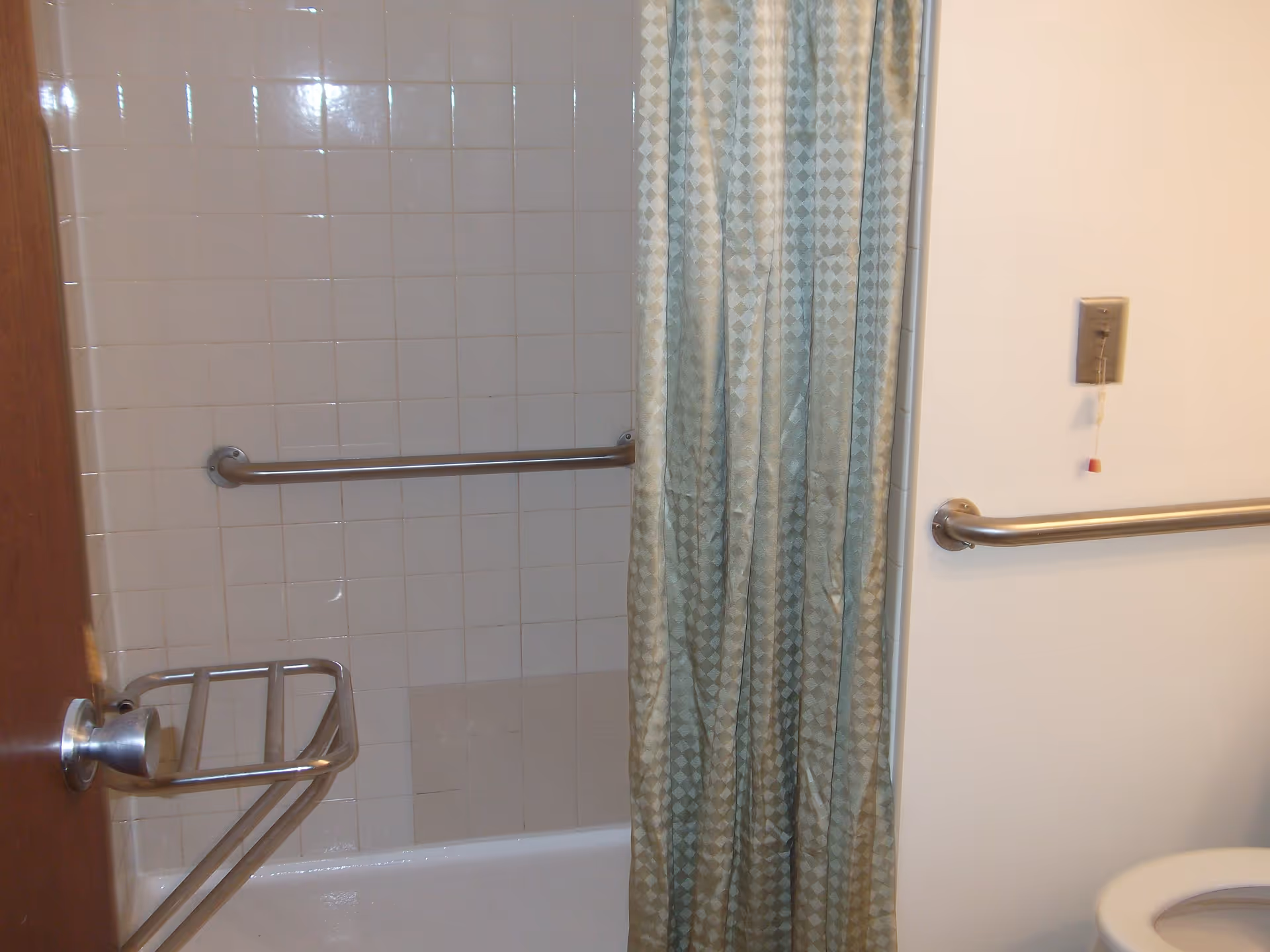 A bathroom with a tiled shower area featuring a metal grab bar and a green patterned shower curtain. There is a metal towel rack attached to the wooden door on the left and another grab bar on the white wall near a toilet. A pull cord hangs from a switch on the wall.