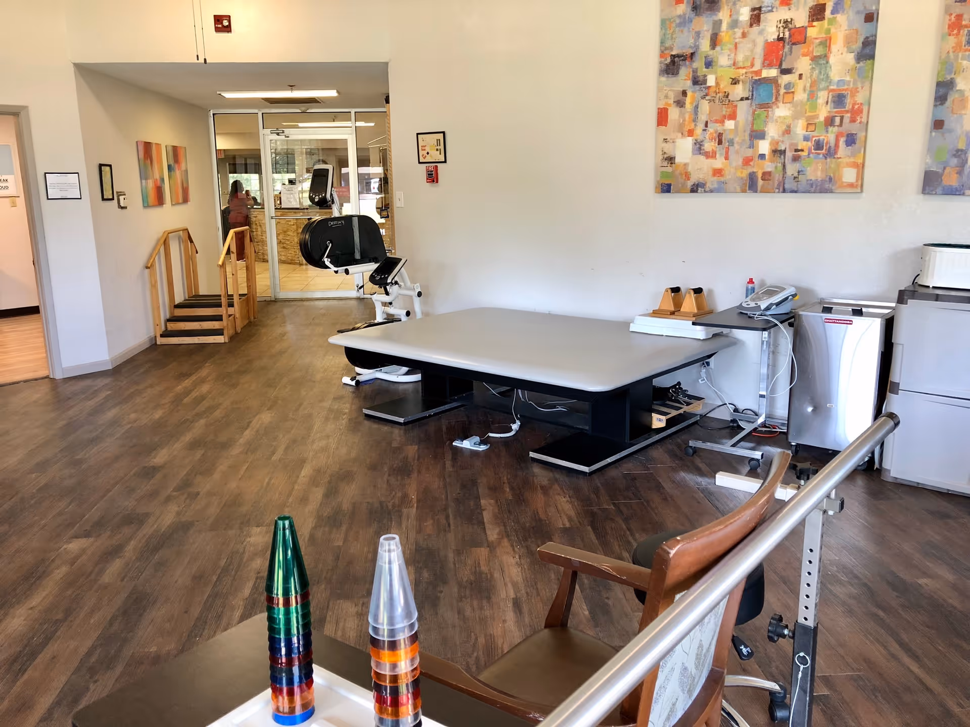 A physical therapy room in a nursing center featuring a large padded therapy table, exercise equipment including a stationary bike, wooden stairs for rehabilitation, and colorful abstract paintings on the walls. The room has wood flooring and a glass door leading to another area.