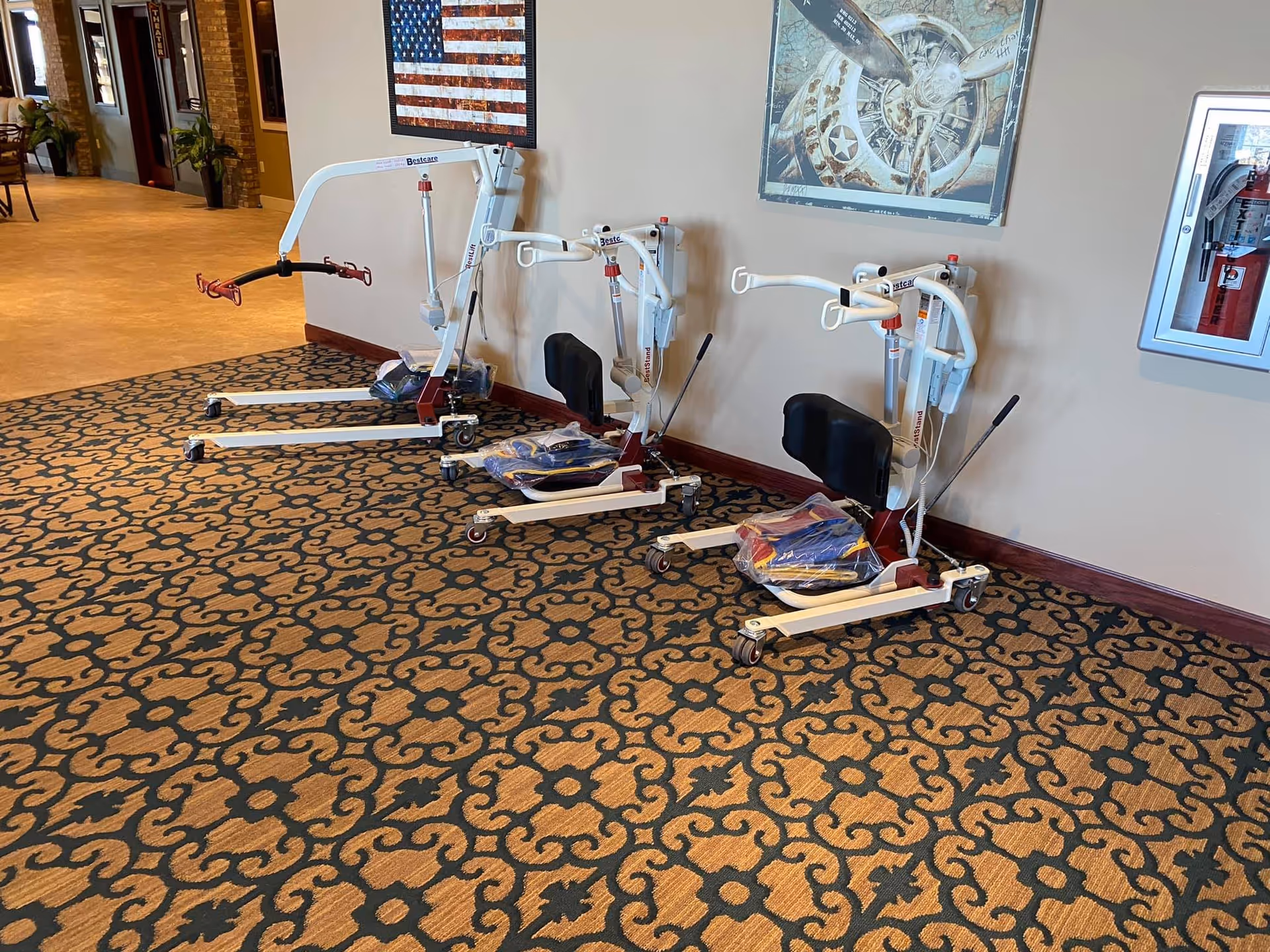 Three white and red patient lifts with black seats are lined up against a beige wall in a hallway with patterned carpet. On the wall above the lifts are two framed pictures, one of an American flag and another of an airplane engine. A fire extinguisher is mounted on the wall to the right.