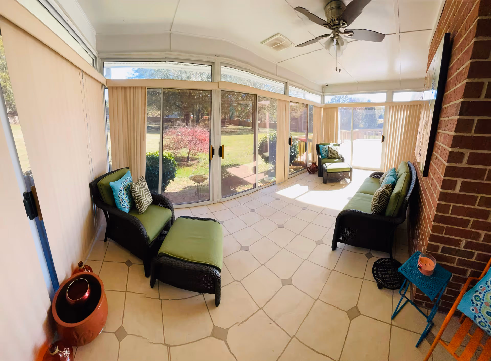A bright sunroom with large sliding glass doors and windows overlooking a green yard. The room has tiled flooring and is furnished with dark wicker seating including a sofa, two armchairs with green cushions, and an ottoman. There are decorative pillows on the chairs and sofa, a ceiling fan above, and a small blue side table with a candle on it. The walls include a brick accent wall and vertical blinds covering the windows.