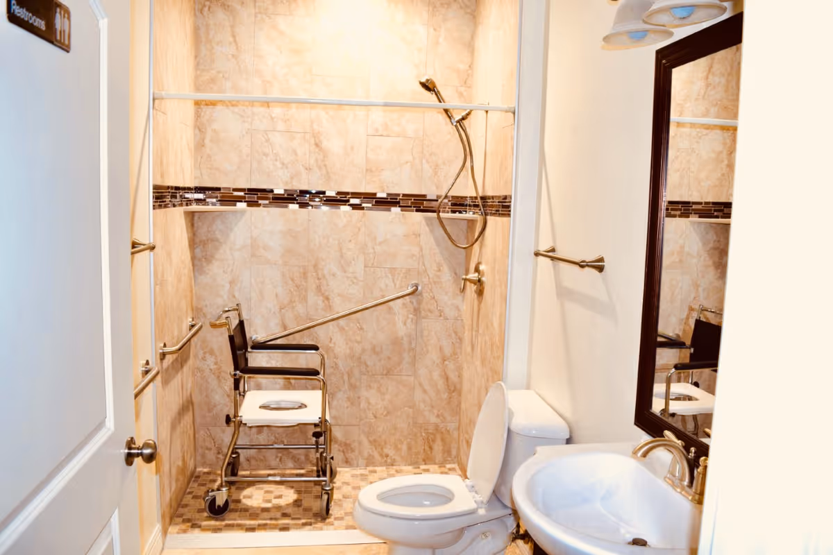 A bathroom with beige tiled walls and floor, featuring a walk-in shower with a handheld showerhead and a shower chair. There are grab bars installed on the walls for support. A toilet with the lid open and a white sink with a mirror above it are also visible. A sign on the door indicates restrooms.
