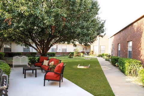Outdoor courtyard area at Morada Waxahachie with a large tree providing shade over a seating area that includes red cushioned chairs and a small table. The courtyard is surrounded by brick and siding buildings with windows, and there is a concrete walkway along the grass.