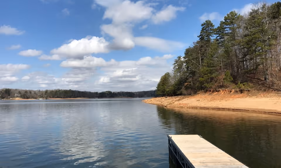 A calm lake with a wooden dock extending into the water on the right side. The shoreline is sandy with a forested area of pine and deciduous trees. The sky is partly cloudy with blue patches visible.