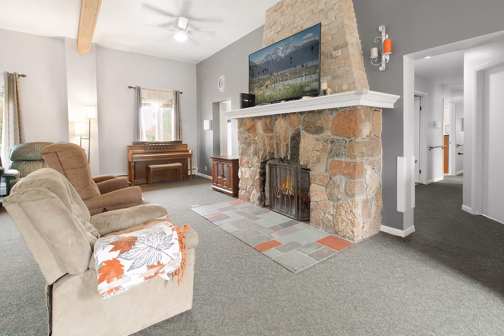 A cozy living room with a stone fireplace and a mounted flat-screen TV above it. There are three recliner chairs, one with a floral blanket draped over the arm. A piano with a bench is positioned against the far wall under a window with curtains. The room has gray walls, carpeted floor, and a ceiling fan with a light. A hallway is visible to the right.