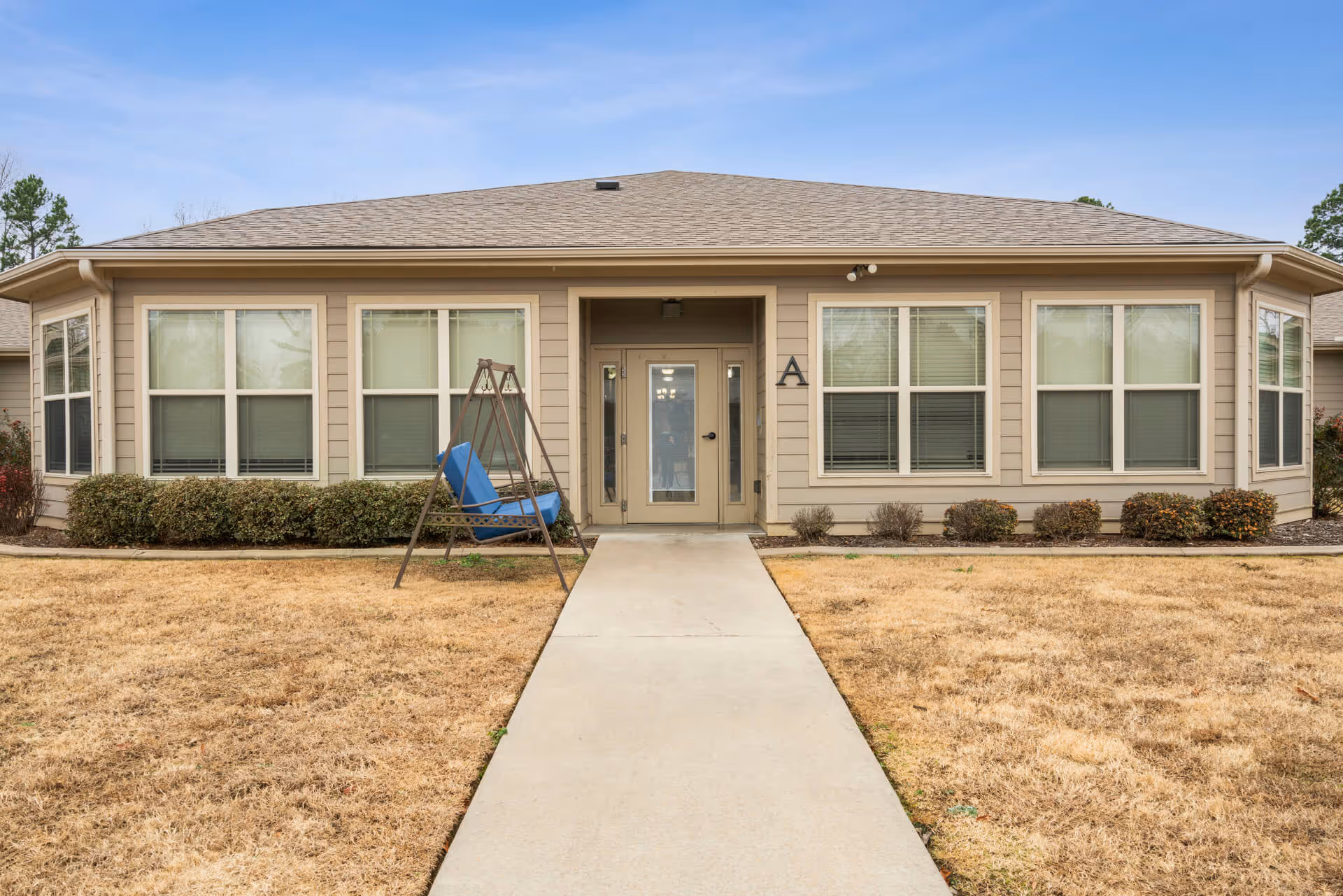 Front exterior view of a single-story building with beige siding and a gray shingled roof. There is a concrete walkway leading to a glass door entrance labeled with the letter 'A'. The building has multiple windows with white frames and blinds. A blue cushioned swing is positioned on the left side of the walkway in front of the building. The surrounding lawn is dry and brown with some bushes near the building.