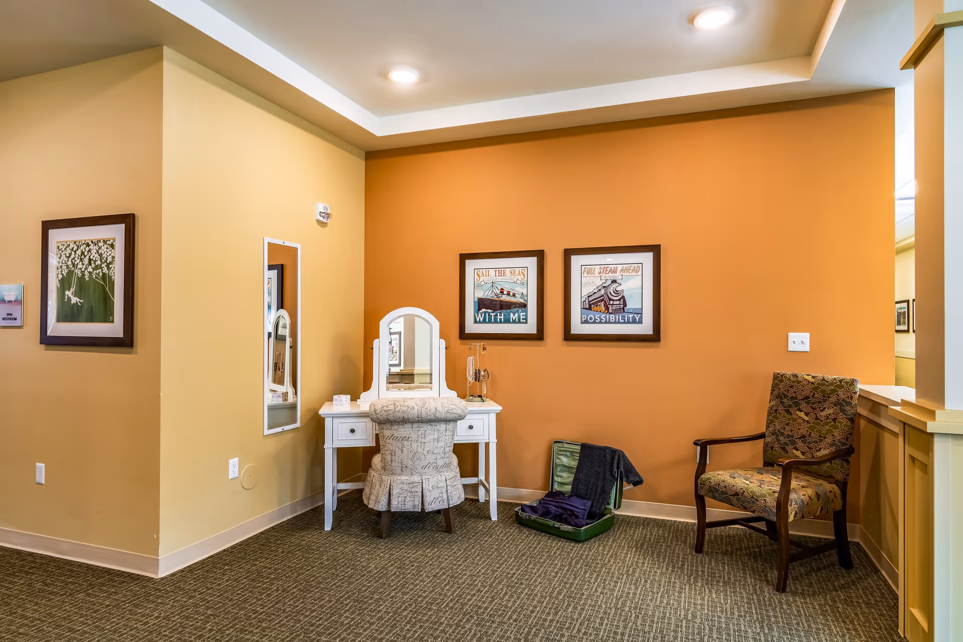 A cozy corner in a senior living facility with a white vanity table and cushioned chair, a patterned armchair, and two framed nautical-themed pictures on an orange accent wall. There is a small open suitcase with clothes on the floor, a mirror on the adjacent wall, and a framed floral artwork on the left wall.